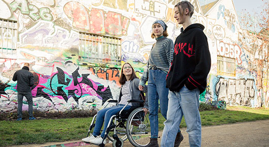 Drei junge Menschen auf einem Weg in einem Park. Im Hintergrund eine Wand mit Graffiti.