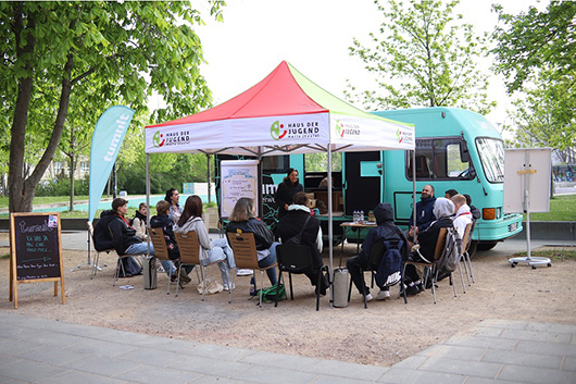 Gruppe junger Menschen vor einem Infobus in einem Kreis sitzend