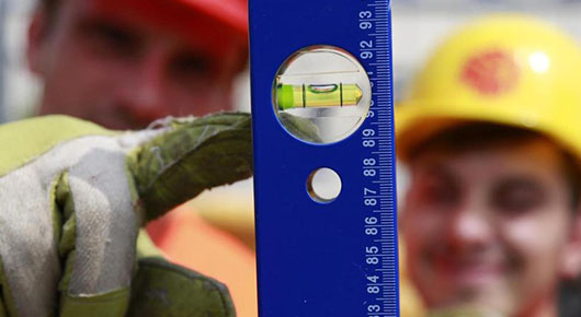 Im Vordergrund eine blaue Wasserwaage, hochkant, auf die ein Finger in einem Arbeitshandschuh zeigt. Im Hintergrund ein Mann mit Schutzhelm.